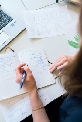 Female designer sketching architectural plans with notes and laptop in a creative workspace.